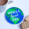 A round glass coaster with an illustration of the world and the words 'World's Best Dad'